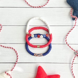 K&ouml;nnte beinhalten: Drei &uuml;bereinanderliegende Armb&auml;nder mit roten, wei&szlig;en und blauen Perlen. Das mittlere Armband hat das Wort "AMERICA" in goldenen Buchstaben auf wei&szlig;en Perlen.