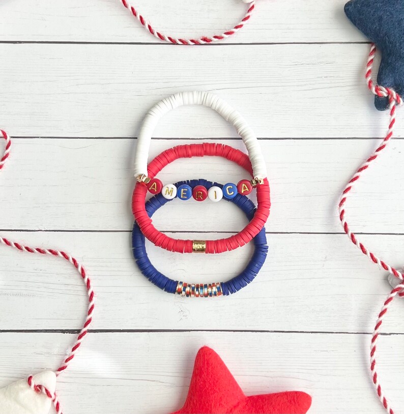 K&ouml;nnte beinhalten: Drei &uuml;bereinanderliegende Armb&auml;nder mit roten, wei&szlig;en und blauen Perlen. Das mittlere Armband hat das Wort "AMERICA" in goldenen Buchstaben auf wei&szlig;en Perlen.