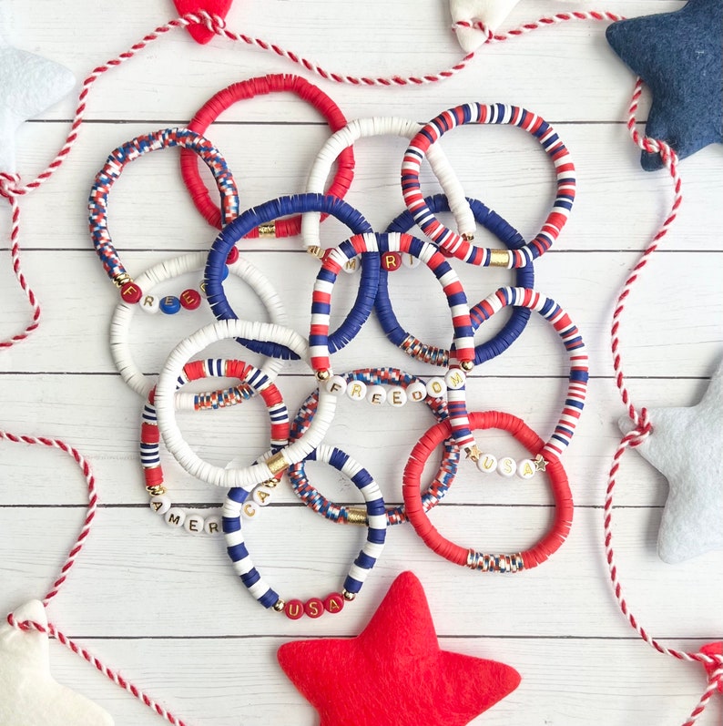 K&ouml;nnte beinhalten: Eine Sammlung von rot-wei&szlig;-blauen Armb&auml;ndern mit goldenen Buchstaben, die "USA", "FREEDOM", "R", "AMER" und "CA" buchstabieren. Die Armb&auml;nder bestehen aus bunten Perlen und sind auf einer wei&szlig;en Holzoberfl&auml;che gestapelt.