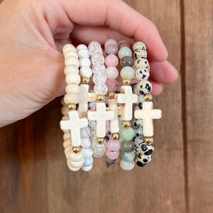 May include: A collection of beaded bracelets in various colors and textures. Each bracelet features a cross charm and gold-colored accents. The beads include white, pink, and speckled designs, creating a layered look.
