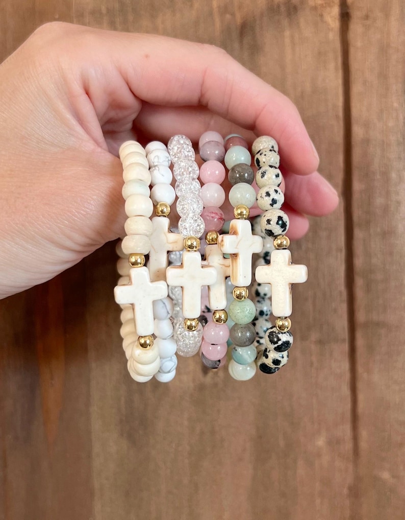 May include: A collection of beaded bracelets in various colors and textures. Each bracelet features a cross charm and gold-colored accents. The beads include white, pink, and speckled designs, creating a layered look.