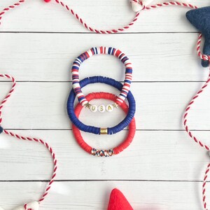 K&ouml;nnte beinhalten: Drei &uuml;bereinanderliegende Armb&auml;nder in den Farben Rot, Wei&szlig; und Blau. Das mittlere Armband hat wei&szlig;e Perlen mit goldenen Buchstaben, die "USA" buchstabieren, und goldenen Sternen.