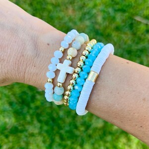 May include: A stack of beaded bracelets in shades of white, gold, and turquoise. One bracelet features a white cross charm. The bracelets are worn on a wrist against a backdrop of green grass.