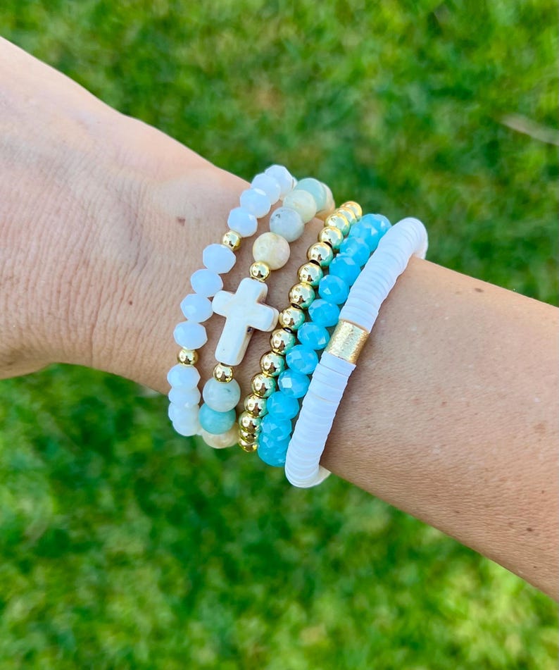 May include: A stack of beaded bracelets in shades of white, gold, and turquoise. One bracelet features a white cross charm. The bracelets are worn on a wrist against a backdrop of green grass.