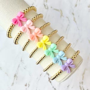 May include: A collection of gold beaded bracelets, each adorned with a colorful bow. The bows are in a rainbow of pastel colors: pink, peach, yellow, green, blue, and purple. The bracelets are displayed on a white, cylindrical surface.