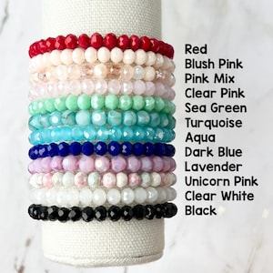 May include: Stack of 12 colorful beaded bracelets in various shades of red, pink, green, blue, white and black. The bracelets are stacked on a white surface.