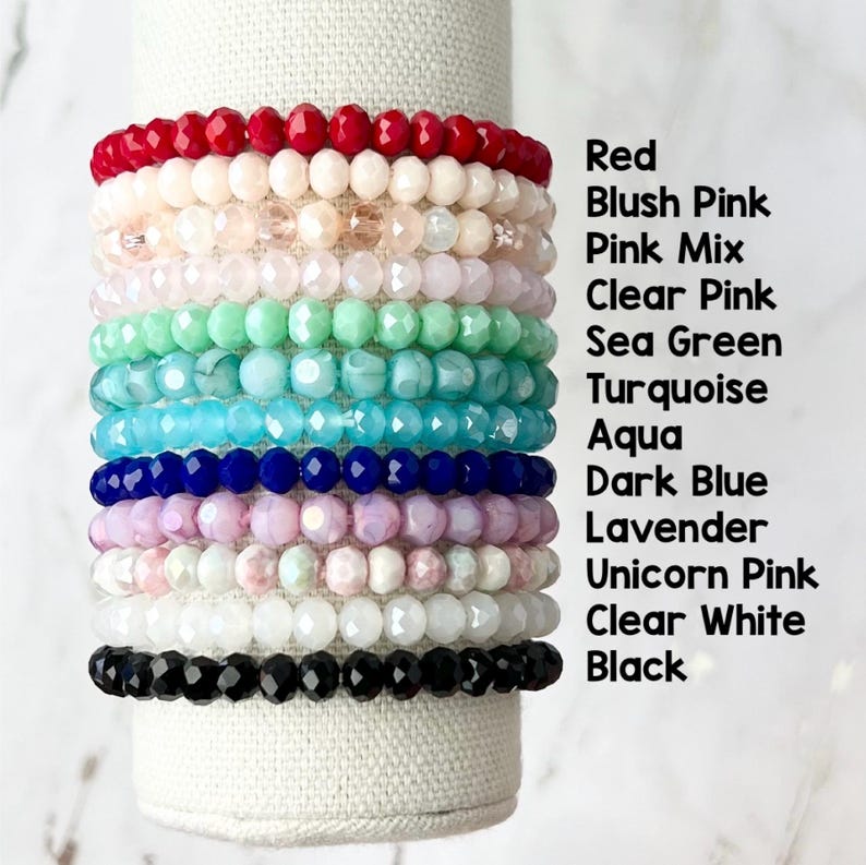 May include: Stack of 12 colorful beaded bracelets in various shades of red, pink, green, blue, white and black. The bracelets are stacked on a white surface.