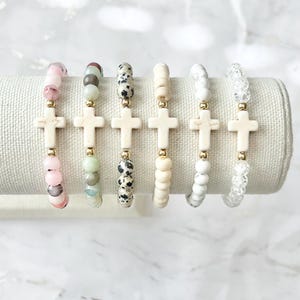 May include: A display of six beaded bracelets with cross pendants. The bracelets feature various colored beads, including pink, green, black and white, and clear. Each bracelet has a cream-colored cross pendant and gold-colored accents.