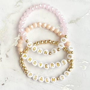 May include: Three beaded bracelets with gold beads and white letter beads spelling out "JOHN 3:16", "PROVERBS", and "PSALM 23". The bracelets are layered on a white surface.