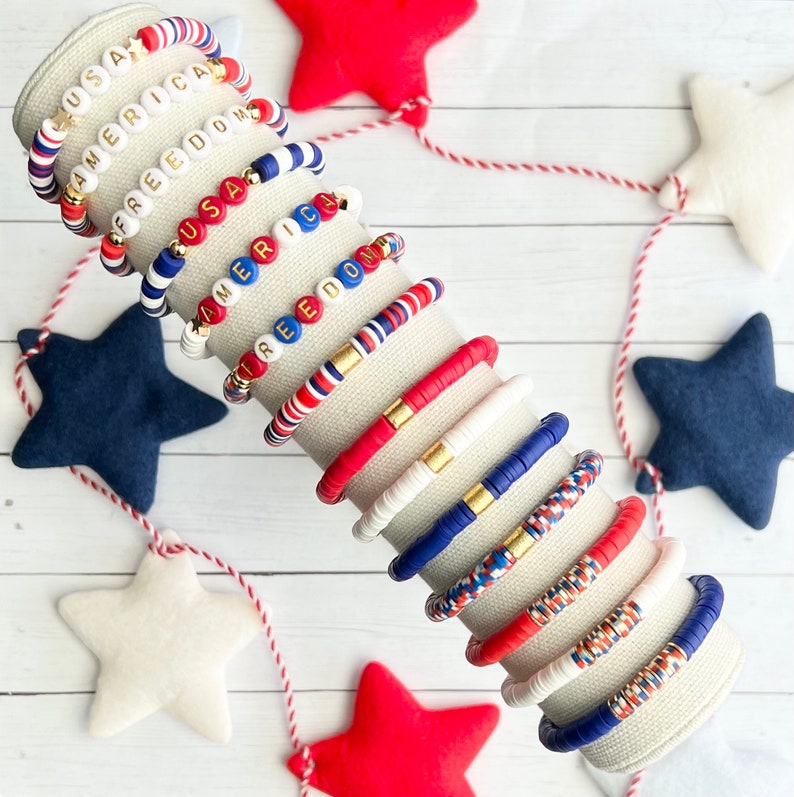 K&ouml;nnte beinhalten: Ein Stapel bunter Perlenarmb&auml;nder mit roten, wei&szlig;en und blauen Perlen. Die Armb&auml;nder buchstabieren die W&ouml;rter "USA", "America", "Freedom", "USA", "America" und "Freedom".