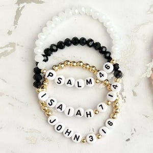 May include: Three beaded bracelets with white beads, black beads, and gold beads. The bracelets have white letter beads spelling out "PSALM 46", "ISAIAH 1", and "JOHN 3:16".