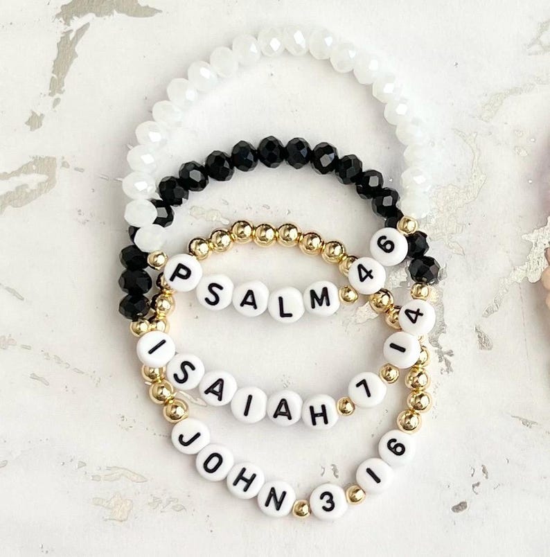 May include: Three beaded bracelets with white beads, black beads, and gold beads. The bracelets have white letter beads spelling out "PSALM 46", "ISAIAH 1", and "JOHN 3:16".