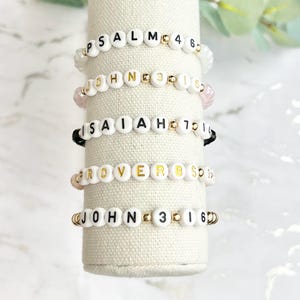 May include: Five white beaded bracelets with gold accents and black lettering spelling out Bible verses: Psalm 46, John 3:16, Isaiah 40:31, Proverbs 3:5, and John 3:16.