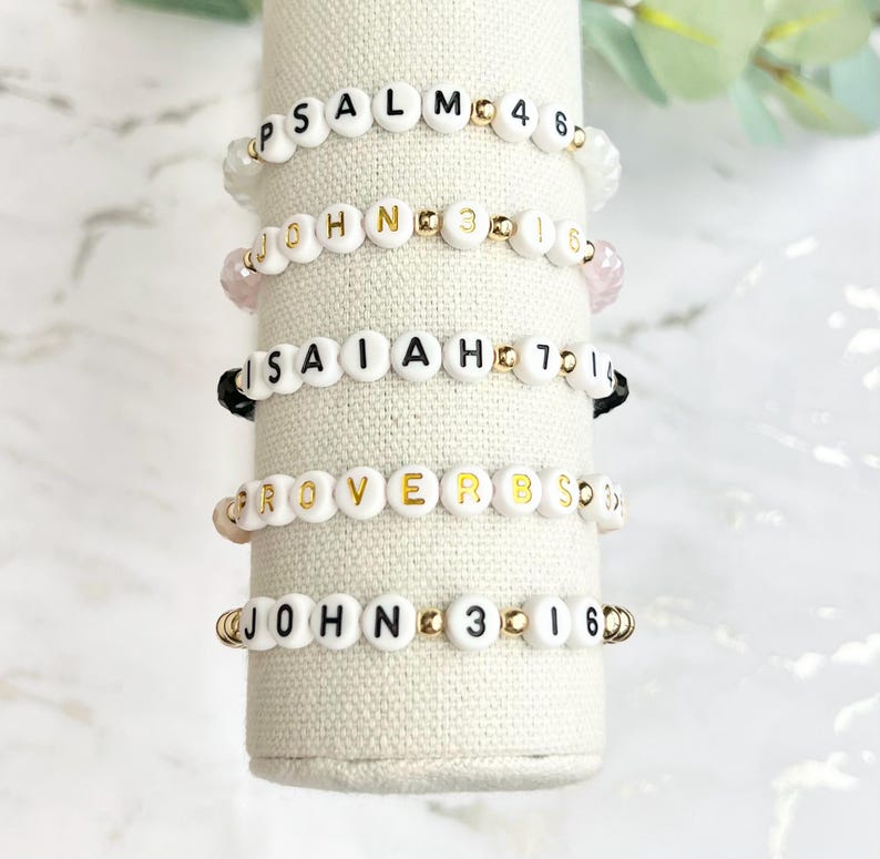 May include: Five white beaded bracelets with gold accents and black lettering spelling out Bible verses: Psalm 46, John 3:16, Isaiah 40:31, Proverbs 3:5, and John 3:16.