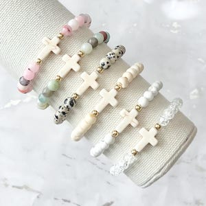 May include: Six beaded bracelets with cross pendants. The bracelets feature various bead colors, including pink, green, black and white. Each bracelet has a cream-colored cross pendant and gold-colored accents. The bracelets are displayed on a white fabric roll.