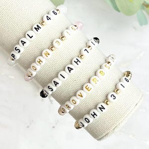 May include: Five white beaded bracelets with gold accents and black lettering. The bracelets spell out "PSALM 4 6", "JOHN 3 16", "ISAIAH 40 31", "PROVERBS 3 5", and "JOHN 3 16".