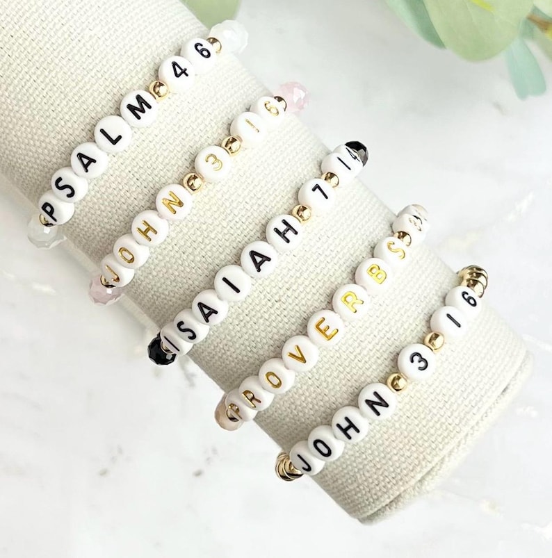 May include: Five white beaded bracelets with gold accents and black lettering. The bracelets spell out "PSALM 4 6", "JOHN 3 16", "ISAIAH 40 31", "PROVERBS 3 5", and "JOHN 3 16".
