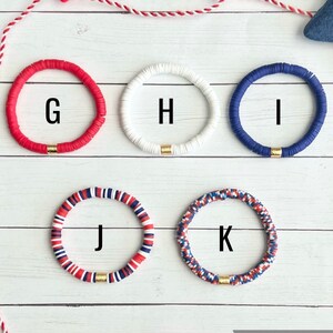 K&ouml;nnte beinhalten: F&uuml;nf bunte Armb&auml;nder mit goldenen Akzenten. Die Armb&auml;nder sind rot, wei&szlig;, blau und eine Kombination aus rot, wei&szlig; und blau. Die Armb&auml;nder sind mit G, H, I, J und K beschriftet.