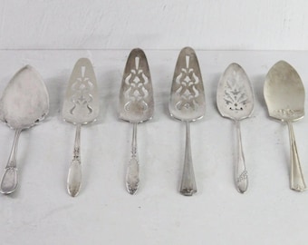 May include: Six vintage silver cake servers with different handle and blade designs. The servers are arranged on a white surface, showcasing their ornate patterns and shapes. The blades have various cut-out designs.