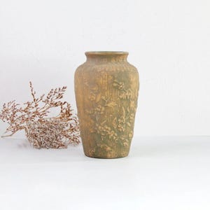 Puede incluir: Un jarrón de cerámica verde con un diseño floral en relieve. El jarrón tiene un cuerpo redondeado y una abertura estrecha.