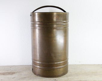 May include: A cylindrical copper container with a dark, weathered patina and a smooth finish. The vessel features a single arched metal handle attached to the top rim and two horizontal embossed bands encircling the body of the container.
