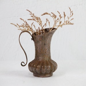 May include: A rustic brown metal pitcher with a unique curved handle. The pitcher is filled with dried flowers.
