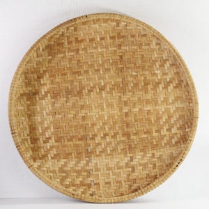 May include: A round, woven basket with a natural, light brown color. The basket has a tight, intricate weave pattern and a slightly raised edge. The basket is likely used for storage or display.