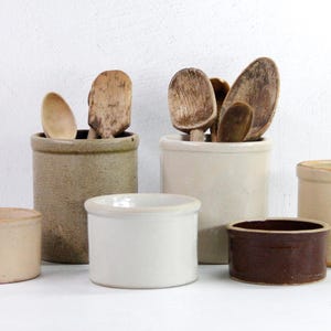 May include: A collection of six vintage ceramic crocks in various sizes and colors, including beige, brown, and white. The crocks are filled with wooden spoons.