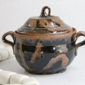 May include: A small, black and brown ceramic pot with a lid and two handles. The pot has a rustic, handmade look.