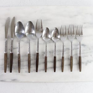 May include: A set of ten stainless steel flatware pieces featuring dark brown wooden handles and silver metal heads. The collection includes two knives, two large spoons, one large fork, two small spoons, and three small forks, all arranged in a row.