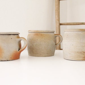 May include: Three ceramic mugs with handles, each with a different glaze. The mugs are a light beige color with brown and white speckles. They are arranged on a white surface.