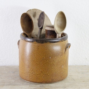 May include: A cylindrical brown stoneware crock with two side handles, filled with several wooden kitchen utensils including spoons and a round flat tool. The crock features a darker brown glazed rim and a speckled textured body finish.