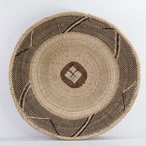 May include: A round, woven basket with a natural beige and brown color scheme. The basket features concentric circles, with a central square design. The outer rim has a textured, dark brown pattern.