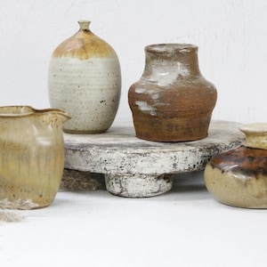 May include: Four ceramic pottery pieces in various shapes and sizes. The pieces are glazed in shades of brown, beige, and white. The pieces are arranged on a white wooden pedestal.