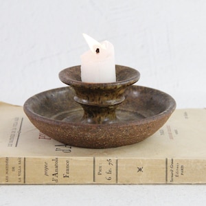 May include: A brown, speckled ceramic candle holder with a wide, shallow base and a central cup, holding a single white pillar candle. The holder features a textured, earthy finish with darker brown mottled spots across the entire surface.