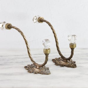 May include: Two wall hooks featuring a twisted brass metal arm and a clear faceted glass knob at each end. The base of each hook is an ornate brass plate with a floral-inspired design, providing a secure mounting point for these hardware pieces.