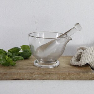 May include: A clear glass mortar and pestle set rests on a wooden cutting board, alongside fresh green basil leaves and a folded beige cloth. The mortar has a rounded bowl and a pouring spout.