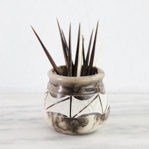 May include: A small, rounded ceramic pot with a marbled grey and white finish, featuring a band of white geometric triangles around the center. The pot is filled with several long, pointed, dark brown and cream-colored wooden sticks.