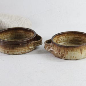 May include: Two ceramic bowls with handles. The bowls have a round shape with a brown rim and a lighter brown base. The handles are small and curved. The bowls are on a white surface.