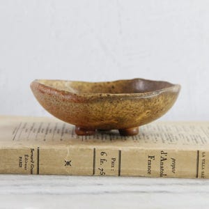 May include: A small, shallow, footed ceramic bowl with a textured, mottled brown and tan glaze. The bowl features an irregular, organic rim shape and rests on three short, rounded feet, showcasing a rustic, earthy appearance and finish.