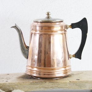 May include: A copper-colored vintage teapot with a silver lid and spout, and a black handle. The teapot is sitting on a wooden surface. The teapot has a classic design and is likely used for brewing tea or coffee.