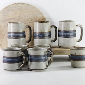 May include: A collection of seven ceramic mugs and one small pitcher, all featuring a speckled off-white glaze with a prominent horizontal band of dark brown and blue stripes around the center. Each piece has a sturdy handle and a cylindrical shape.