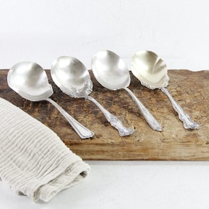 May include: Four silver-toned metal spoons with wide, rounded bowls and decorative embossed handles. The spoons are arranged in a row, each featuring a distinct pattern on the handle, with a light-colored, textured fabric cloth positioned to the left.