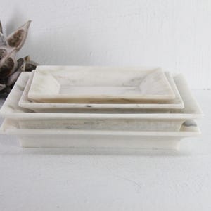 May include: A stack of three rectangular marble trays in varying sizes. The trays have a light, creamy color with subtle veining. The edges are slightly raised, creating a shallow basin. A dried seed pod is in the background.