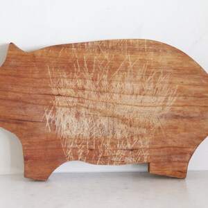 May include: A wooden cutting board in the shape of a pig. The board is a warm brown color with visible wood grain and numerous knife marks in the center. The board has a small notch for a leg.