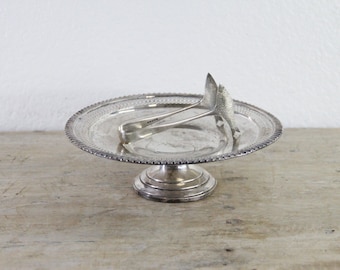 May include: A silver-toned pedestal dish with a circular, perforated rim and a tiered base. Resting inside the dish is a pair of silver-toned metal tongs featuring a textured, triangular gripping end and a smooth, curved handle design.
