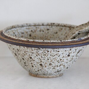 May include: A white ceramic bowl with brown speckles and a brown rim. The bowl has a smaller bowl inside, also with brown speckles and a brown rim. The bowl is sitting on a white surface.