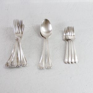 May include: A collection of vintage silverware, including forks and a spoon, arranged on a light gray surface. The forks are grouped in sets, and the spoon is positioned between them. The silverware has a shiny, reflective surface.
