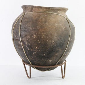 May include: A rustic, round, brown pottery vase with a narrow neck, secured by a metal wire cage. The vase sits on a simple, rusty metal stand. The aged appearance suggests a vintage or antique piece.
