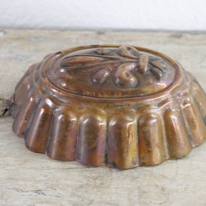 May include: An oval copper mold with a fluted side design and a central embossed floral motif. The metallic surface shows a weathered patina with dark brown and orange tones. A small circular metal hanging loop is attached to one side.